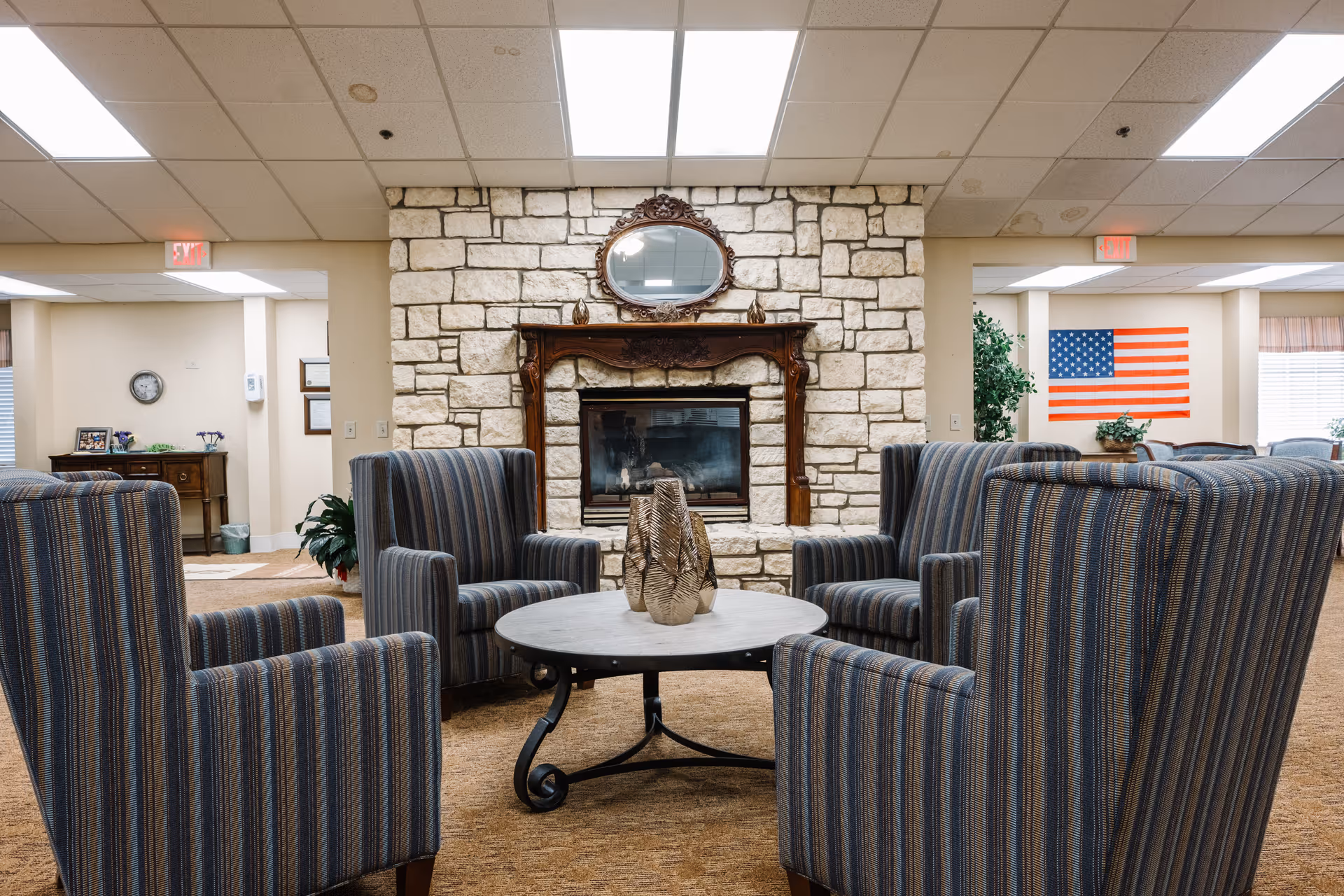 A cozy seating area in a senior living facility featuring four striped armchairs arranged around a round coffee table with decorative vases. Behind the seating area is a stone fireplace with a wooden mantel and an ornate oval mirror above it. The room has a beige carpet and ceiling tiles, with an American flag visible on the wall in the background.