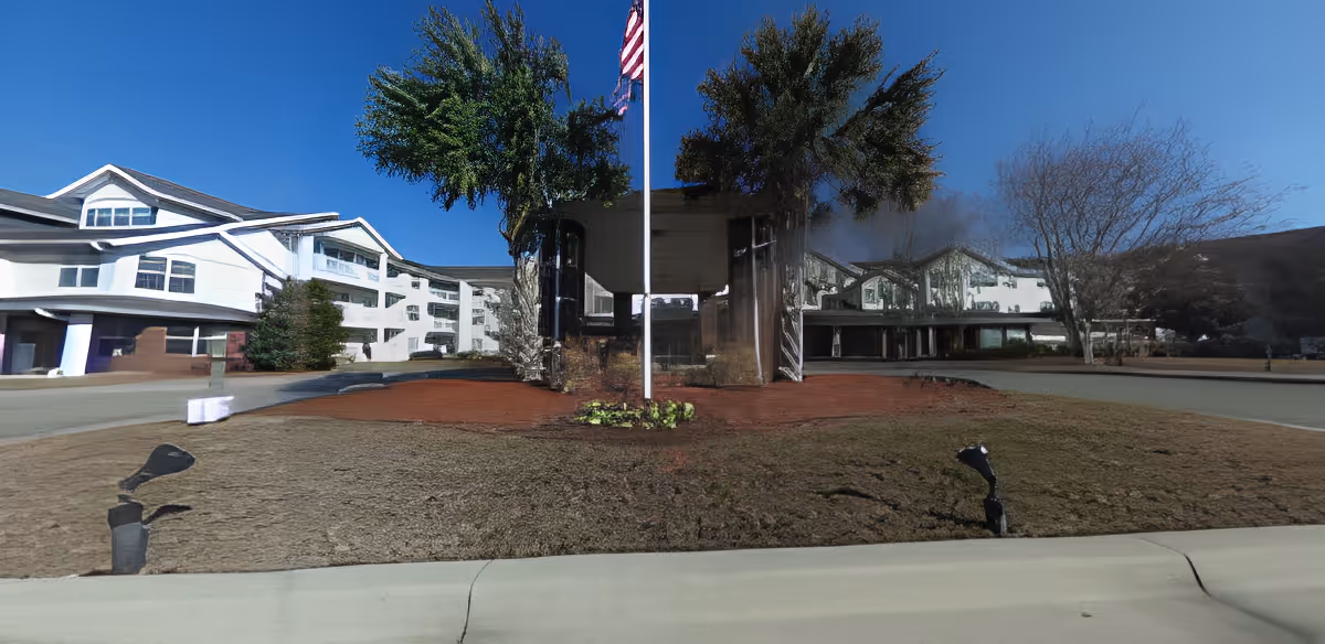 Front exterior view of a large senior living facility building with white walls and multiple windows, a flagpole with an American flag in the center, some trees, and a clear blue sky.