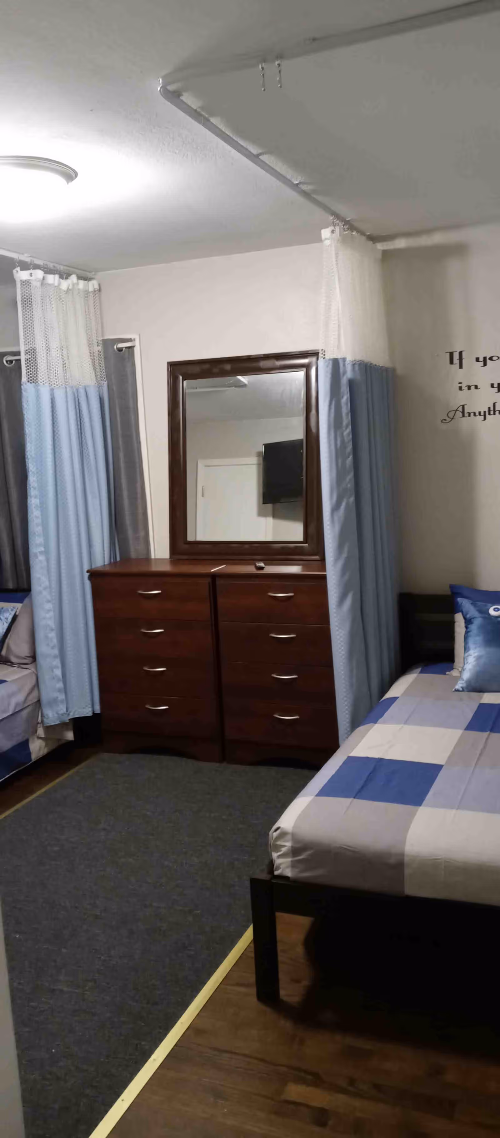 A bedroom with two single beds, each with blue and white checkered bedding. Between the beds is a dark wooden dresser with a large mirror above it. Light blue curtains hang around the beds, and a gray carpet covers part of the wooden floor. A wall decal with partially visible text is on the right wall.
