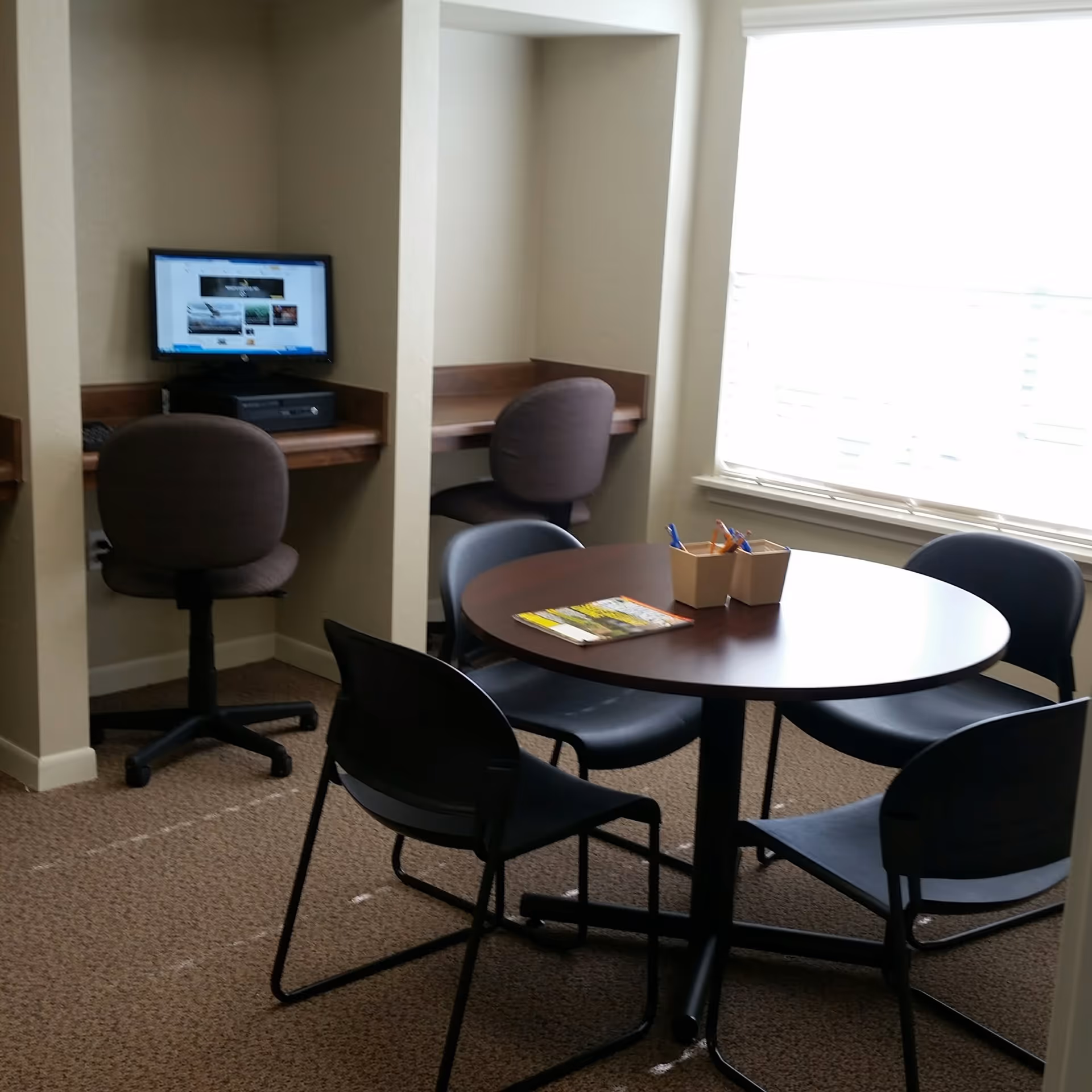 Small communal room with a round table and four chairs near a window and two built-in computer workstations.