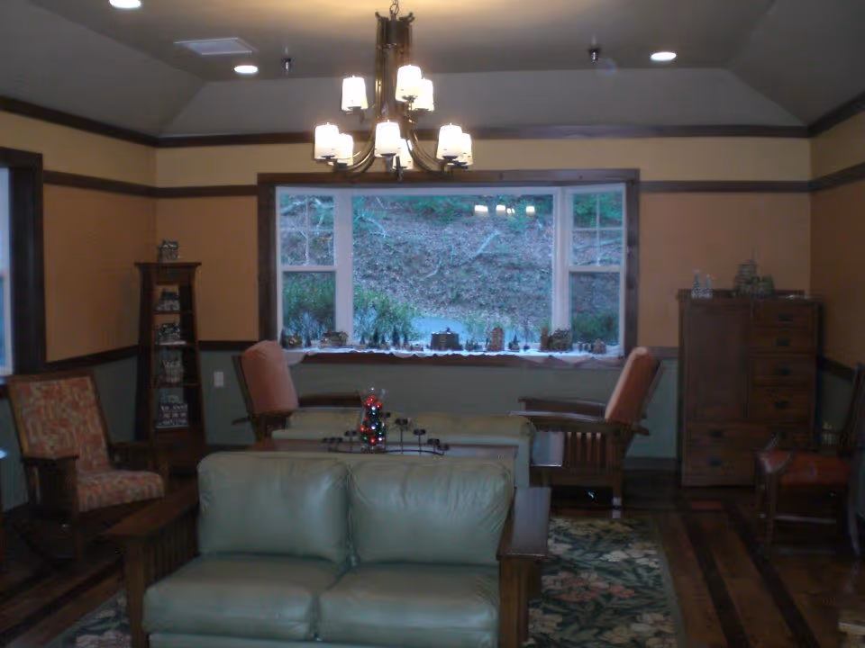 A cozy living room with a green leather sofa in the foreground, two armchairs with orange cushions, a wooden chest of drawers, and a tall narrow shelf. A large window with a view of trees and a decorative display on the windowsill is centered on the back wall. A chandelier with multiple lights hangs from the ceiling, and a floral rug covers part of the wooden floor.