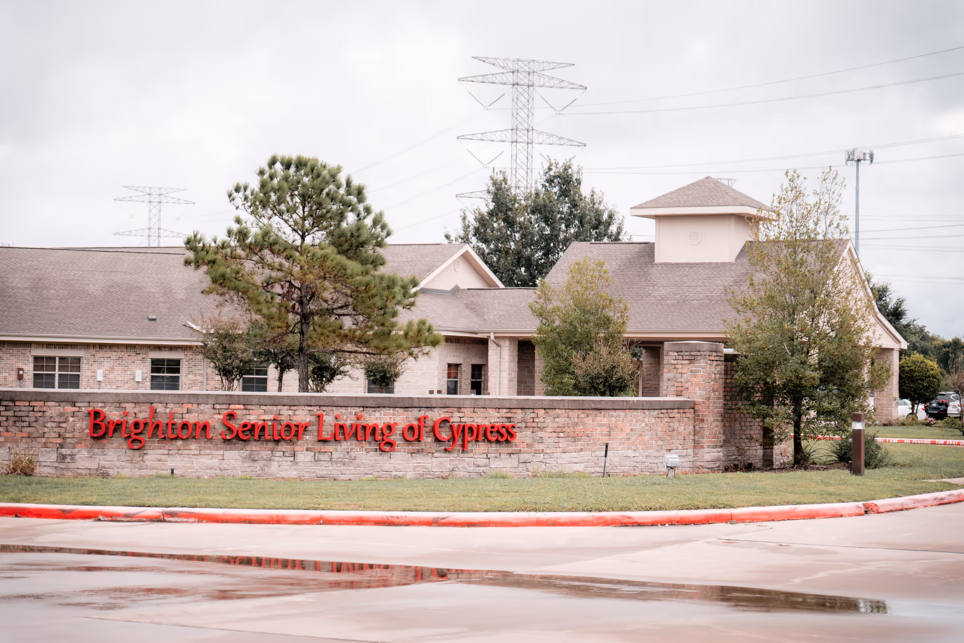 Exterior view of Brighton Senior Living of Cypress facility showing a brick wall with the facility's name in red letters, surrounded by trees and a building with a sloped roof in the background under a cloudy sky.