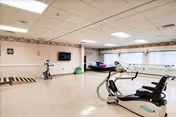 A spacious rehabilitation room with exercise equipment including a stationary bike and a treadmill. The room has beige walls with a floral border near the ceiling, a wall-mounted television, large windows with white curtains, and a hospital bed in the background.