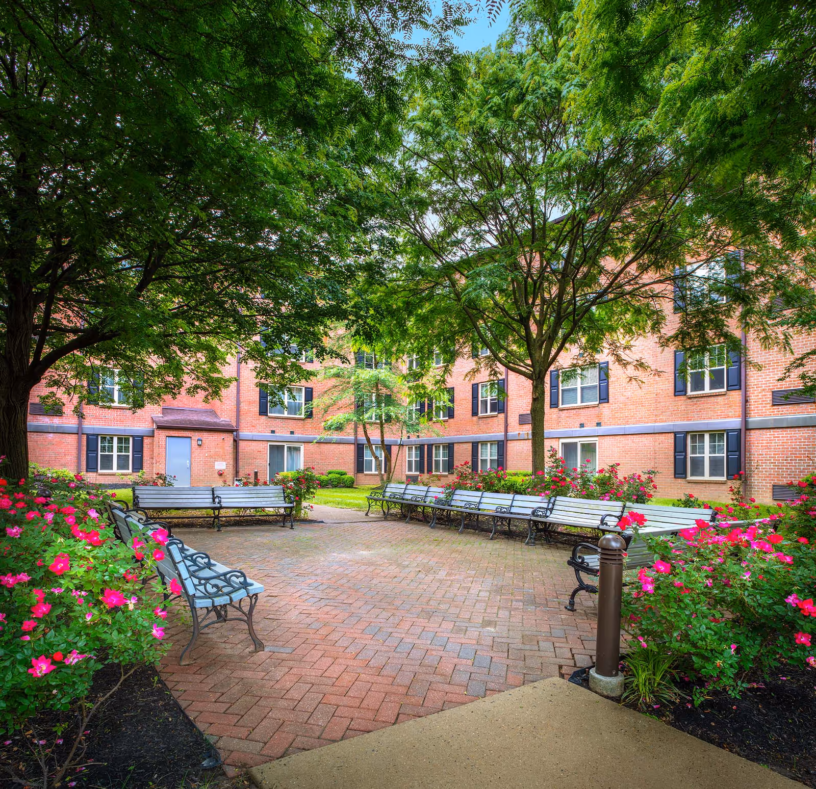 A tree-shaded brick courtyard with benches, flowering shrubs, and a surrounding red-brick residential building.
