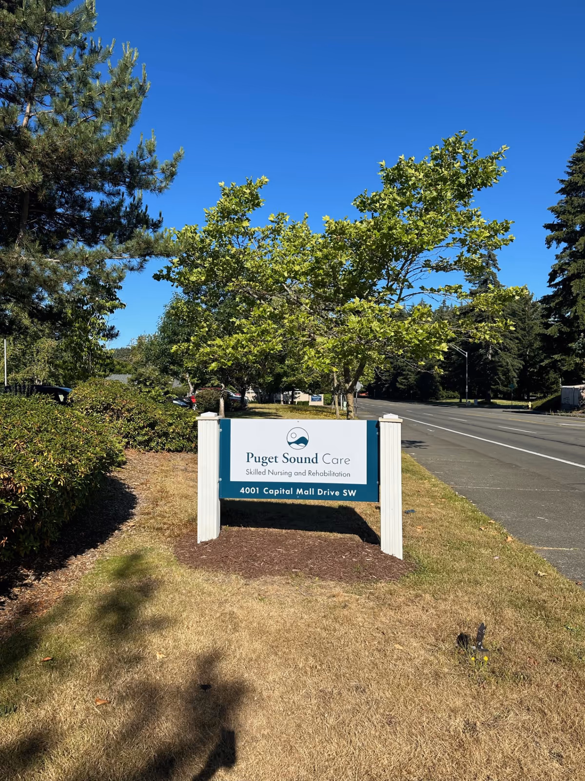 Outdoor view of a sign for Puget Sound Care Skilled Nursing and Rehabilitation located at 4001 Capital Mall Drive SW, surrounded by green bushes and trees under a clear blue sky.