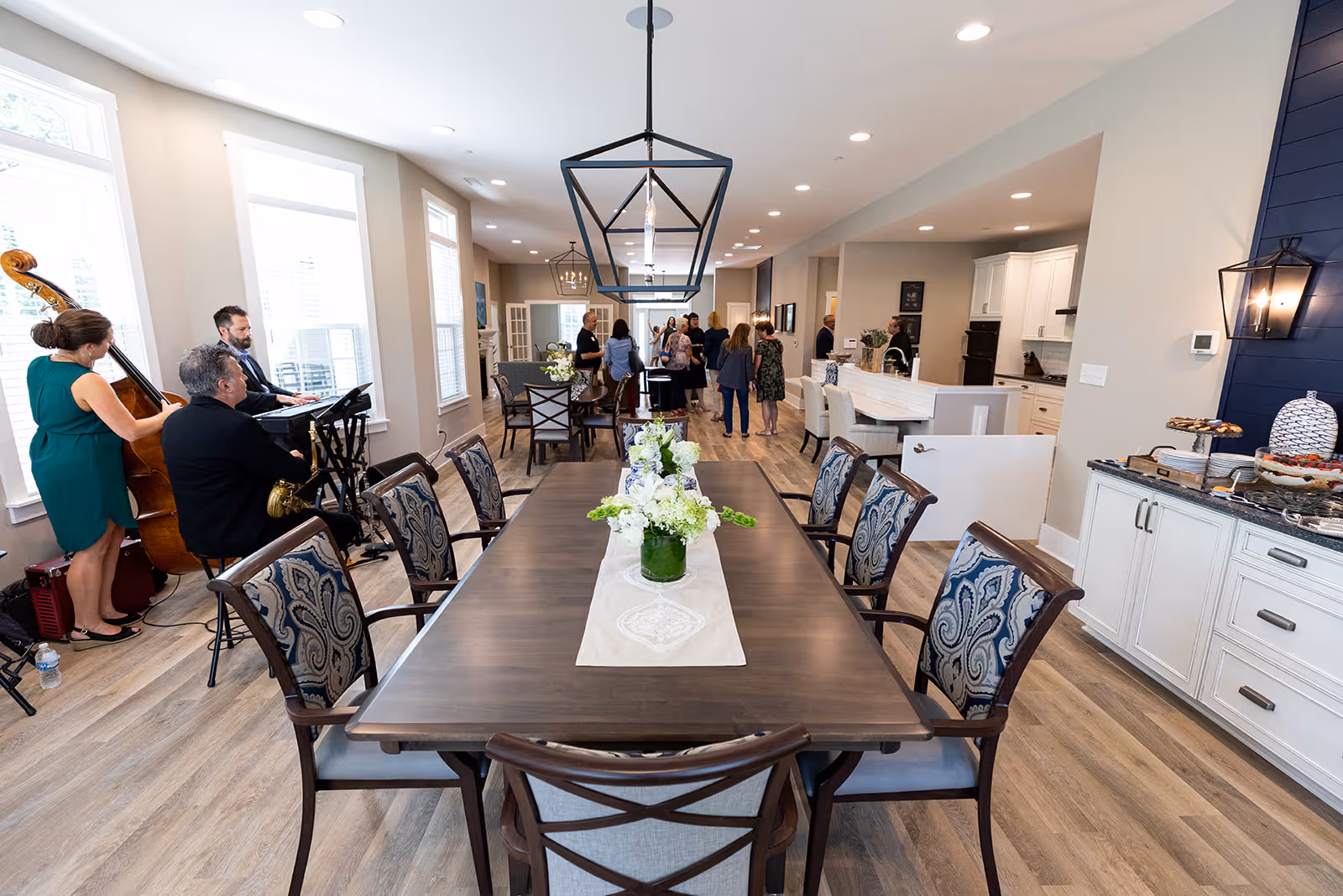 A spacious dining area in a senior living facility with a long wooden dining table surrounded by patterned chairs. The table is decorated with a white runner and floral centerpieces. To the left, a small live band is playing music near large windows. In the background, several people are gathered and conversing near a kitchen area with white cabinetry and a breakfast bar. The room has modern lighting fixtures and wood flooring.