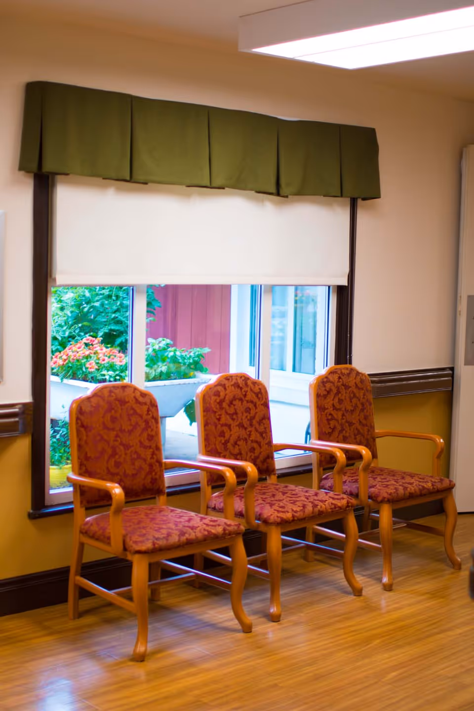 Three wooden chairs with red patterned upholstery are lined up in front of a window with a green valance and a white roller shade. Outside the window, there are plants and flowers visible.