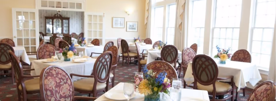 Bright dining room with round tables set with white tablecloths, floral centerpieces, and upholstered chairs by large windows.
