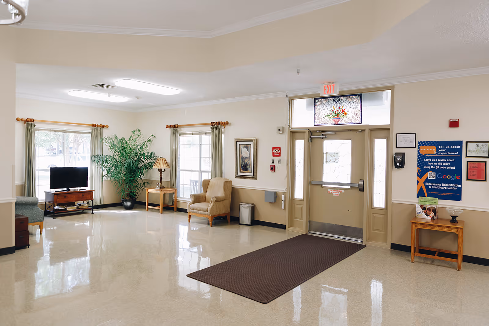 A bright and clean common area in a senior living facility with large windows covered by green curtains, a TV on a wooden stand, a potted plant, two armchairs, a small wooden table with a lamp, and an exit door with decorative glass panels. There are informational posters and a hand sanitizer dispenser on the wall near the door.
