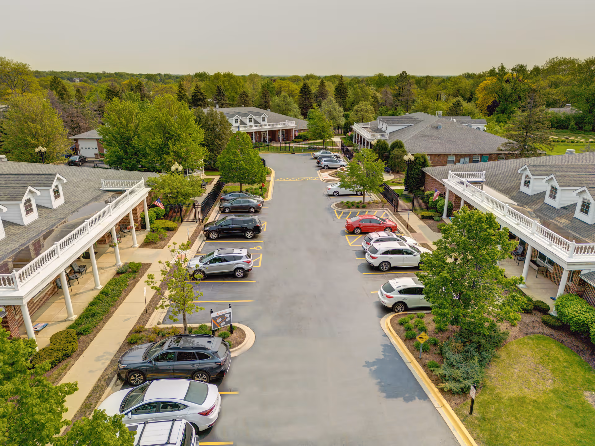 Aerial view of a senior living facility parking lot with several parked cars, surrounded by buildings with white railings and columns. The area is landscaped with green trees and bushes, and there is a dense forest in the background under a clear sky.