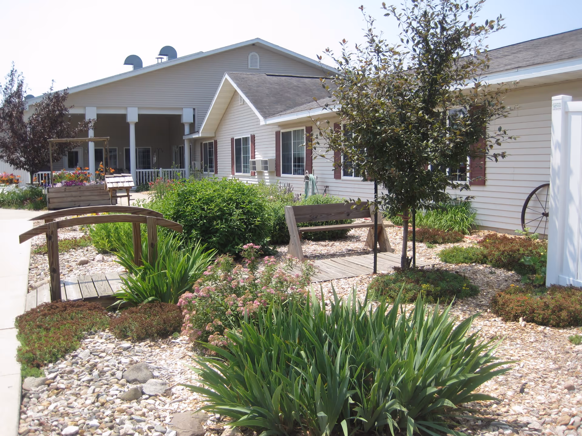 Outdoor garden area at Arlington Place at Grundy Center featuring a small wooden footbridge, a wooden bench, various green plants, shrubs, and flowers, with a beige building with red shutters in the background under a clear sky.