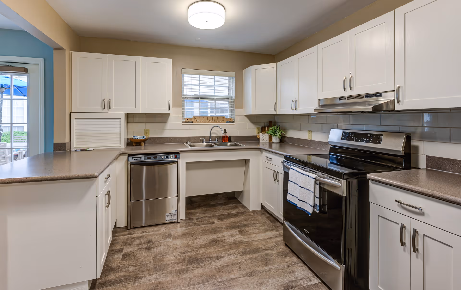 A clean and modern kitchen with white cabinets, stainless steel dishwasher and oven, a double sink under a window with blinds, and a light fixture on the ceiling. The countertops are brown, and the floor has a wood-like finish. There is a small decorative sign that says 'KITCHEN' on the windowsill.
