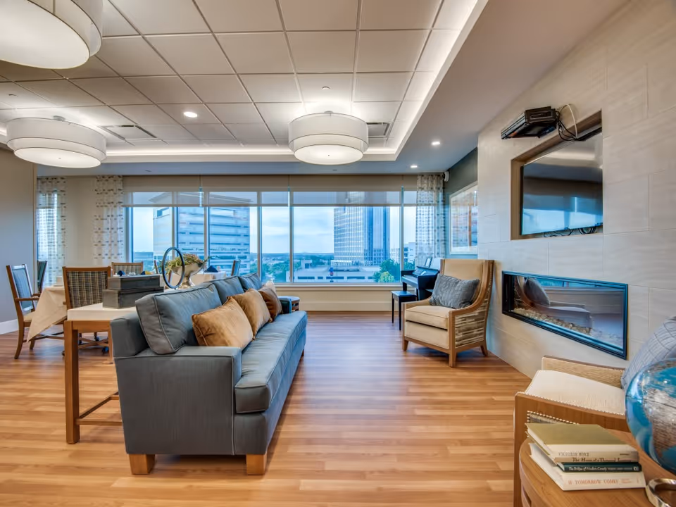 A bright and spacious living room area with a large window offering a cityscape view. The room features a blue sofa with brown cushions, two beige armchairs, a modern built-in fireplace, a wall-mounted TV, and a wooden floor. In the background, there is a dining table with chairs near the window.