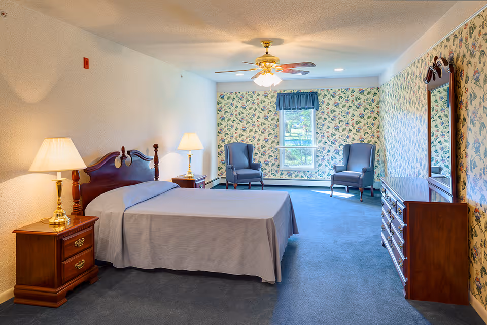 Spacious traditional bedroom with a bed, wooden furniture, two armchairs by a window, and floral wallpaper.