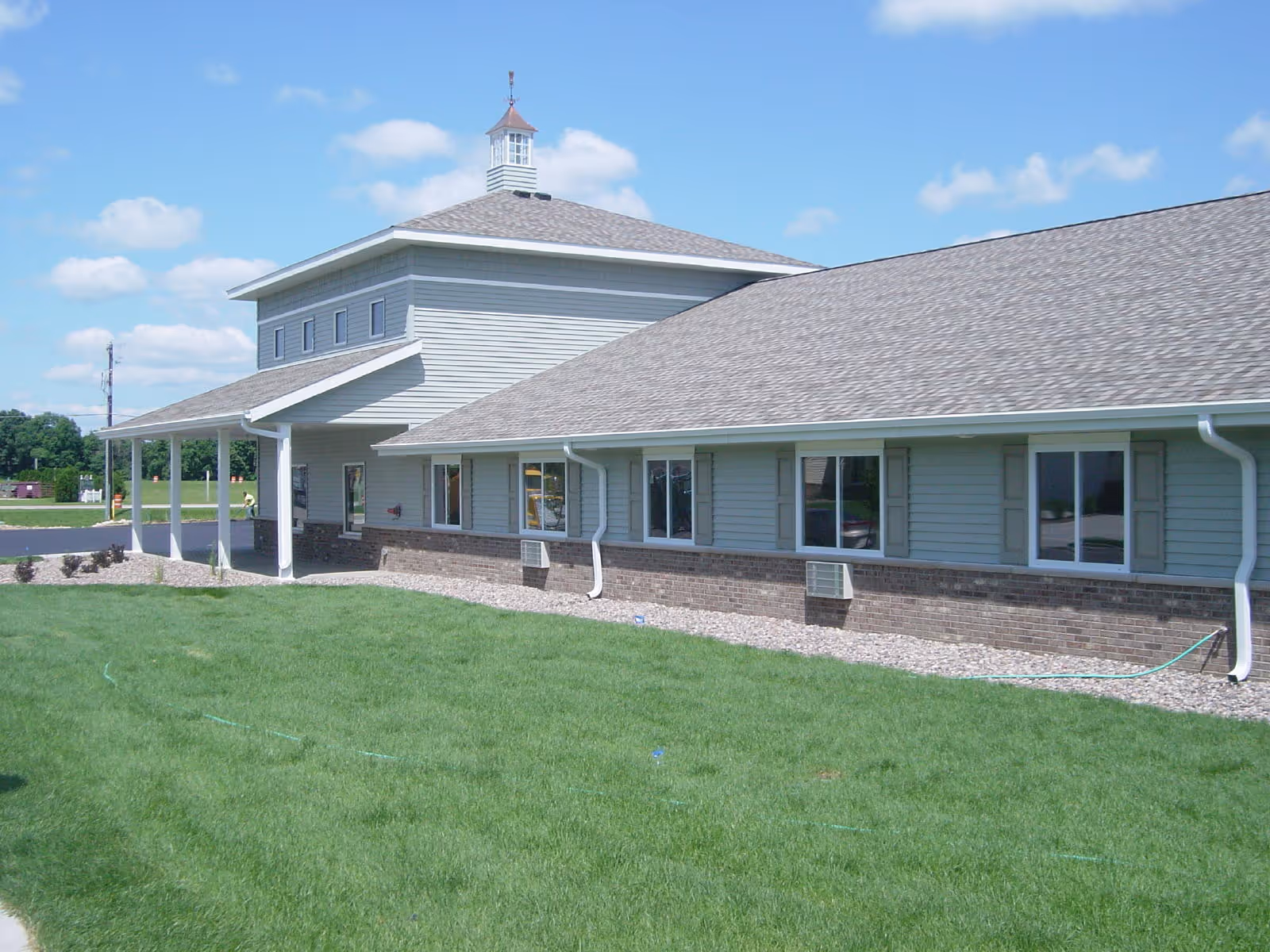 Exterior view of a single-story building with light gray siding and a brick base, featuring multiple windows and a covered entrance. The building is surrounded by a well-maintained green lawn under a blue sky with some clouds.