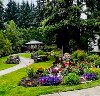 A landscaped garden area with colorful flowers, green shrubs, and trees surrounding a paved walkway leading to a gazebo in the background.