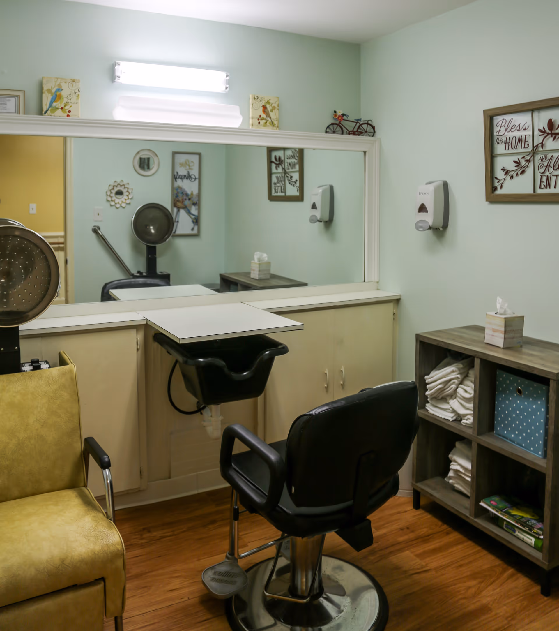 Interior of a small salon area with a black salon chair in front of a large mirror, a yellow chair to the left, a hair dryer, and a wooden shelf with folded towels and a tissue box. The walls are light green with framed decorative signs and a soap dispenser mounted on the wall.