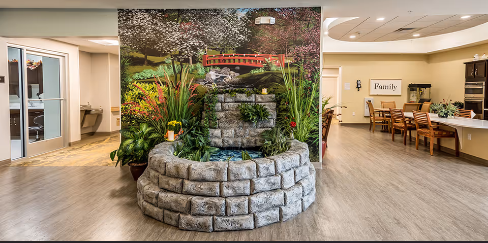 Indoor area of a senior living facility featuring a decorative stone water fountain with plants in front of a mural depicting a garden scene with a red bridge and trees. To the right, there is a dining area with wooden chairs and tables, a sign on the wall that says 'Family', and kitchen appliances. To the left, there are glass doors leading to another room and a water fountain on the wall.