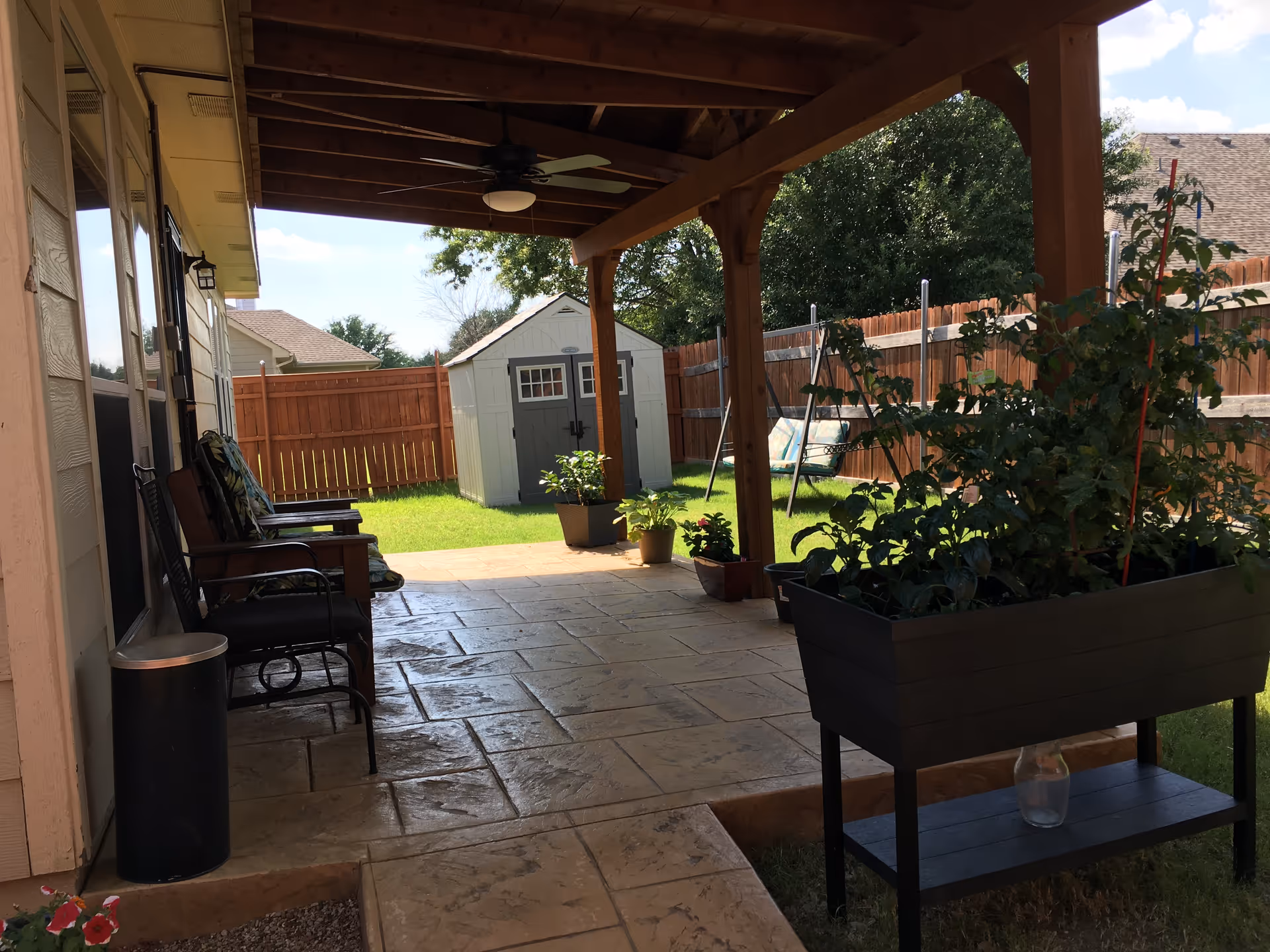 Covered patio area with wooden beams and ceiling fan, featuring chairs with cushions along the wall, potted plants, a raised garden bed, and a small shed in the fenced backyard with green grass and a swing set.