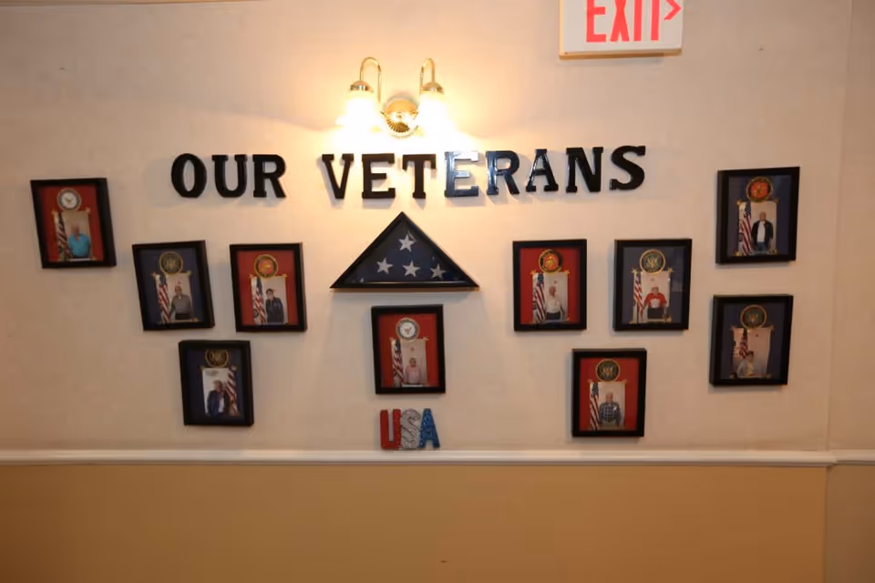A hallway interior wall display reading 'OUR VETERANS' with framed photos, a folded American flag, and a 'USA' decoration under a light and an exit sign above.
