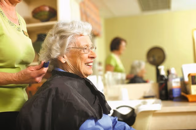 An elderly woman with white hair and glasses is smiling while getting her hair styled by a caregiver in a salon or hairdressing area. The caregiver is standing behind her, and the background shows a mirror and salon equipment.