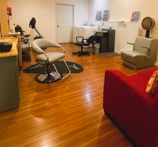 Interior view of a hair salon area with wooden flooring, featuring salon chairs, a hair washing station, hair dryer, and a red couch in the foreground.