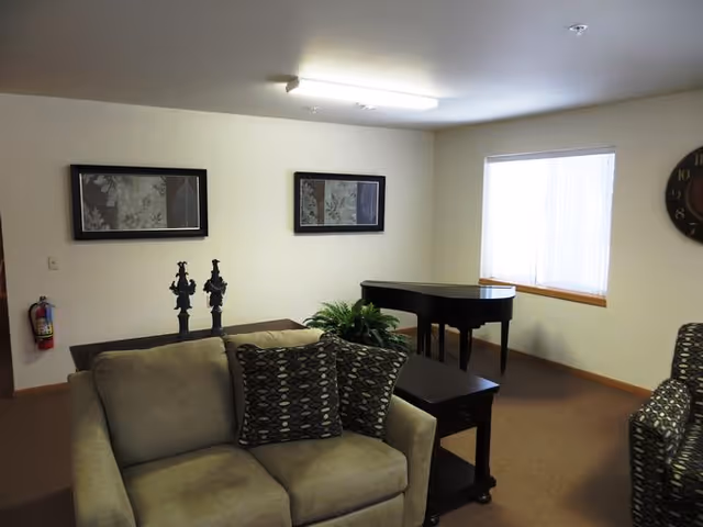 A cozy living room area with a beige loveseat adorned with two patterned pillows, a dark wooden side table, a grand piano near a window with vertical blinds, two framed artworks on the wall, and a fire extinguisher mounted on the wall.