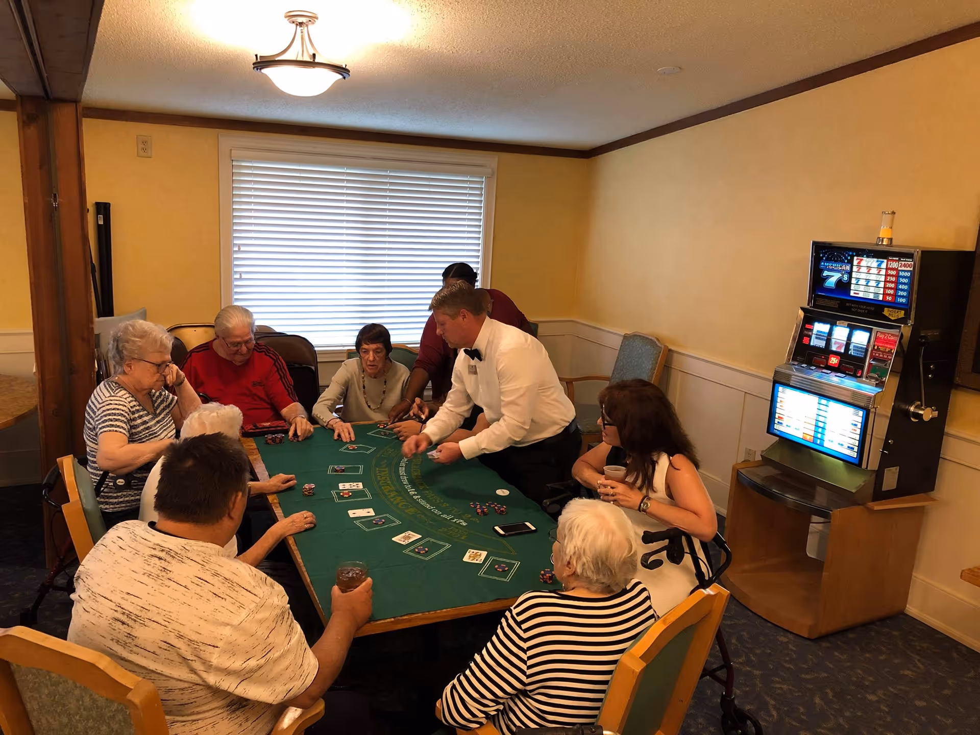 A group of elderly people sitting around a green felt card table playing a card game with a dealer in a white shirt and black bow tie. The room has beige walls, a window with blinds, and a slot machine in the corner. Some players have drinks and chips on the table.