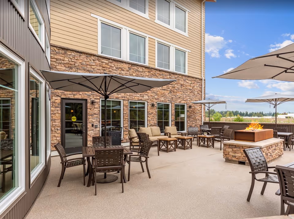 Outdoor patio with tables, chairs, umbrellas, cushioned seating and a stone fire pit alongside a multi-story building.