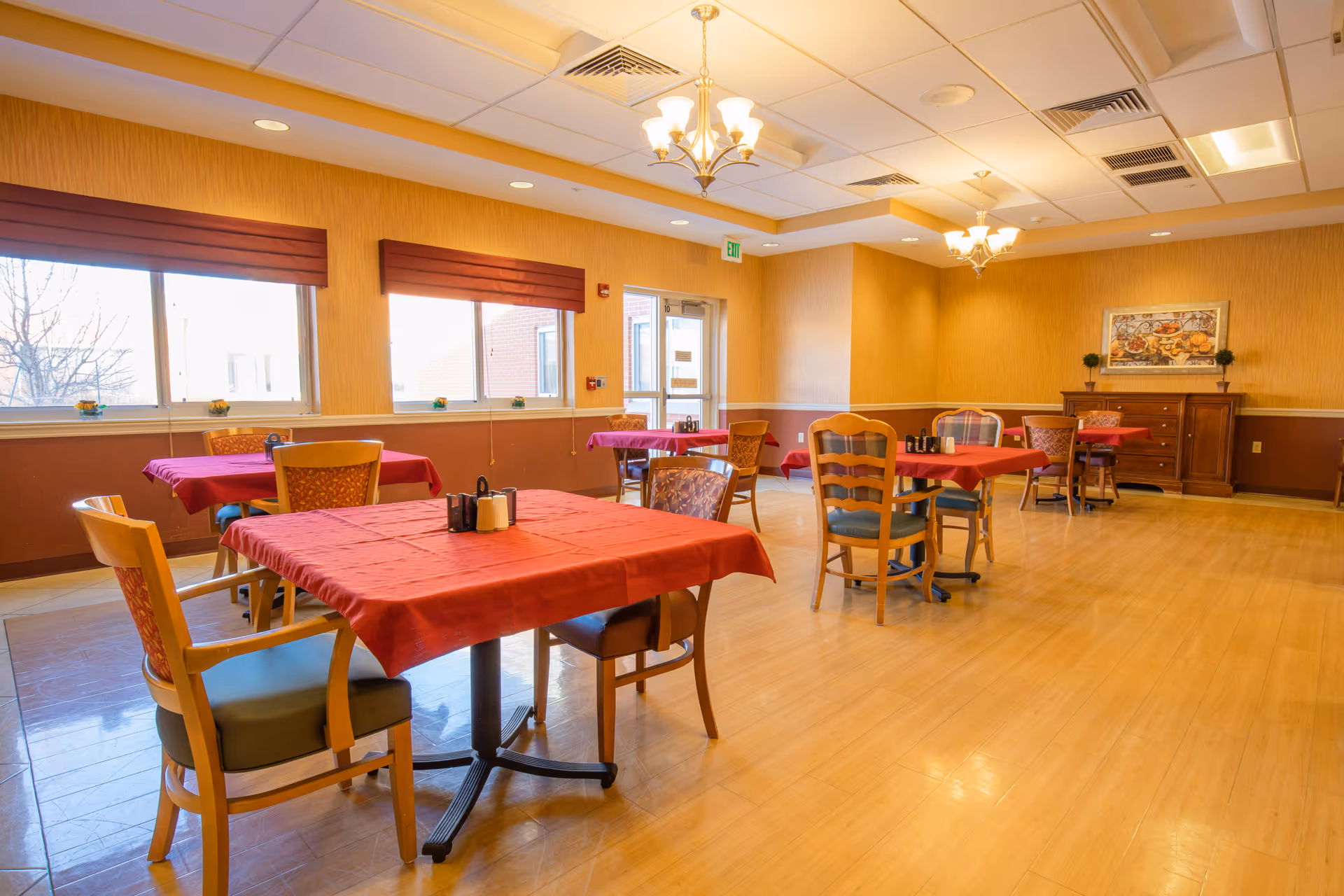 A dining room with several tables covered in red tablecloths, each surrounded by wooden chairs with cushions. The room has large windows with red valances, warm yellow walls, and wooden flooring. There are chandeliers hanging from the ceiling and a wooden sideboard with a framed painting on the far wall.