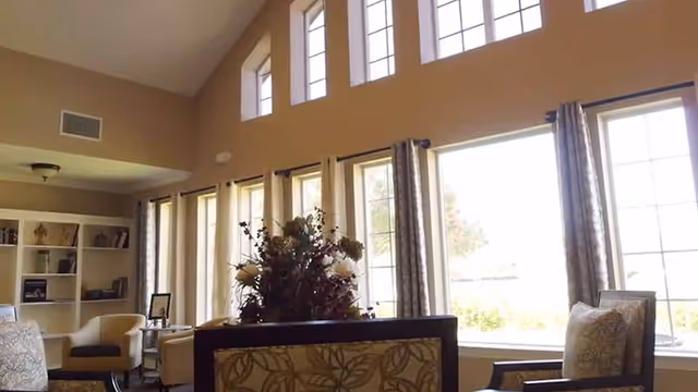 A bright living room area with large windows letting in natural light. The room features multiple chairs with patterned upholstery, a floral arrangement on a table, and built-in shelves with decorative items. Light-colored walls and curtains frame the windows, creating a warm and inviting atmosphere.