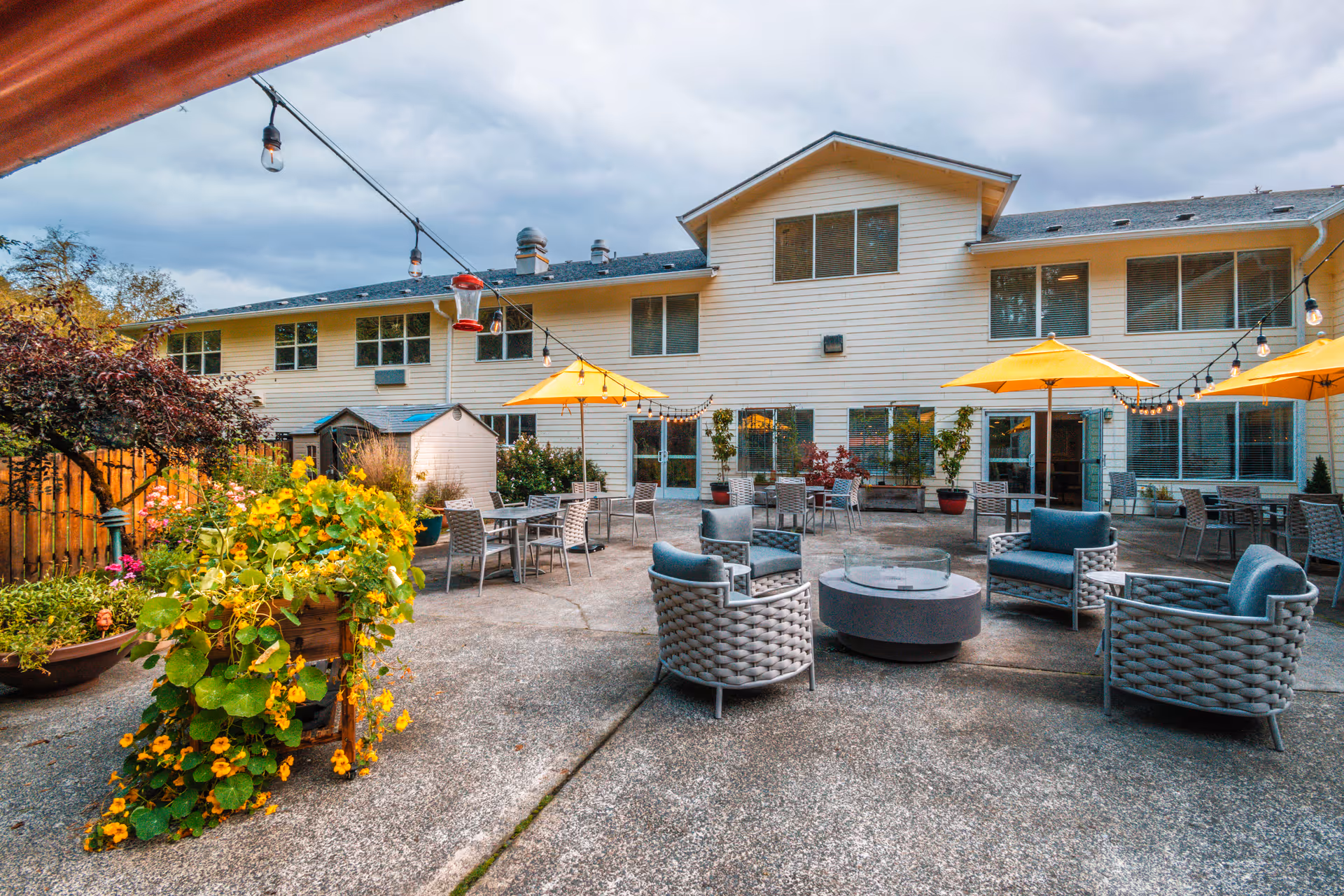 Outdoor courtyard with patio seating, umbrellas, string lights and planters in front of a two-story assisted living building.