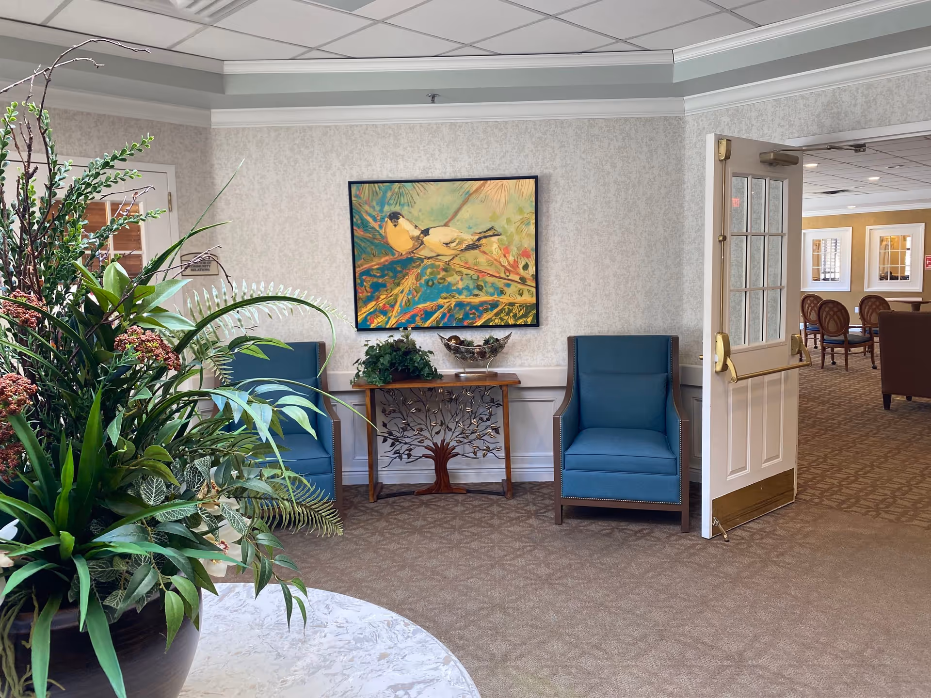 A senior living common area with two blue armchairs flanking a decorative console table, a large potted plant in the foreground, and an open door to another lounge.