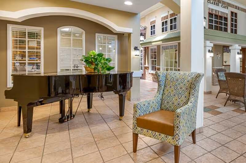 Interior view of a senior living facility featuring a black grand piano with a green plant on top, a patterned armchair with a brown seat cushion, tiled flooring, and a cafe area named 'Clares Cafe' in the background with tables and chairs.