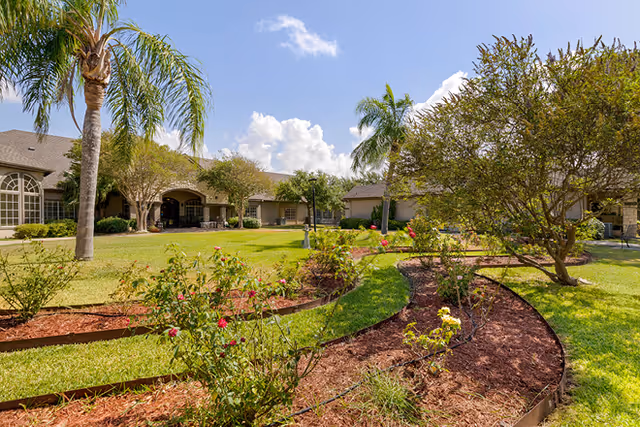 Landscaped courtyard with rose beds, palm trees, and a single-story senior living building under a blue sky.