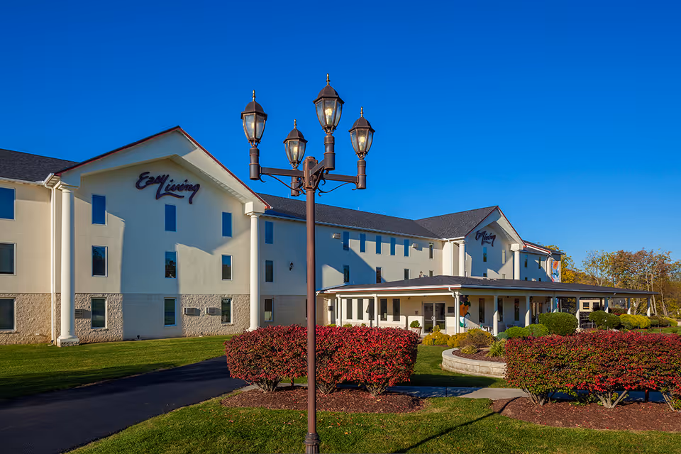 Front exterior of the Easy Living senior living building with landscaped shrubs, a covered entrance, and a decorative lamp post under a clear blue sky.