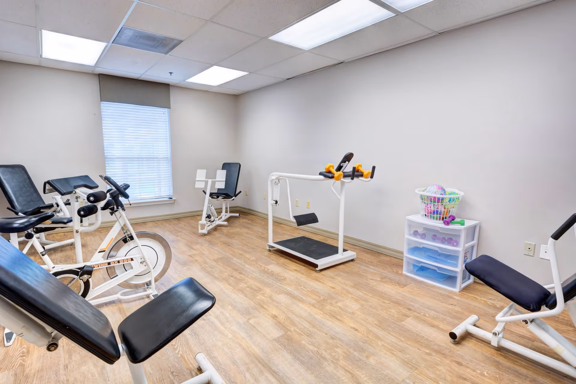 A small exercise room with several pieces of fitness equipment including stationary bikes, a treadmill, and weight machines. There is a window with blinds, light-colored walls, and wood flooring. A plastic drawer unit with a basket of colorful balls and small dumbbells is also visible.