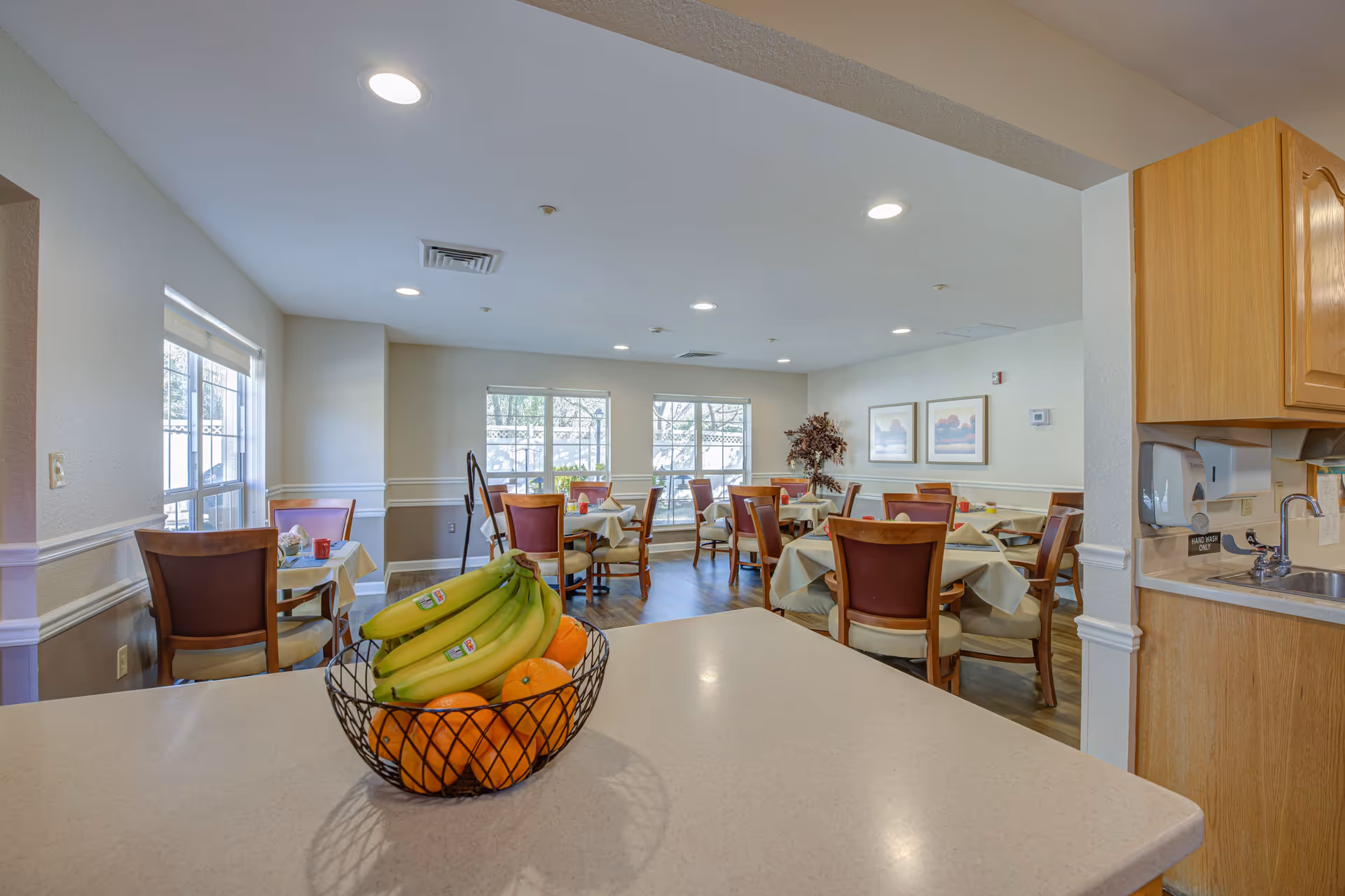 Interior view of a dining room in a senior living facility with multiple tables covered with light-colored tablecloths and wooden chairs with cushioned seats. A basket of bananas and oranges is placed on a countertop in the foreground. The room has large windows letting in natural light, framed artwork on the walls, and a small sink area with hand wash dispenser on the right.