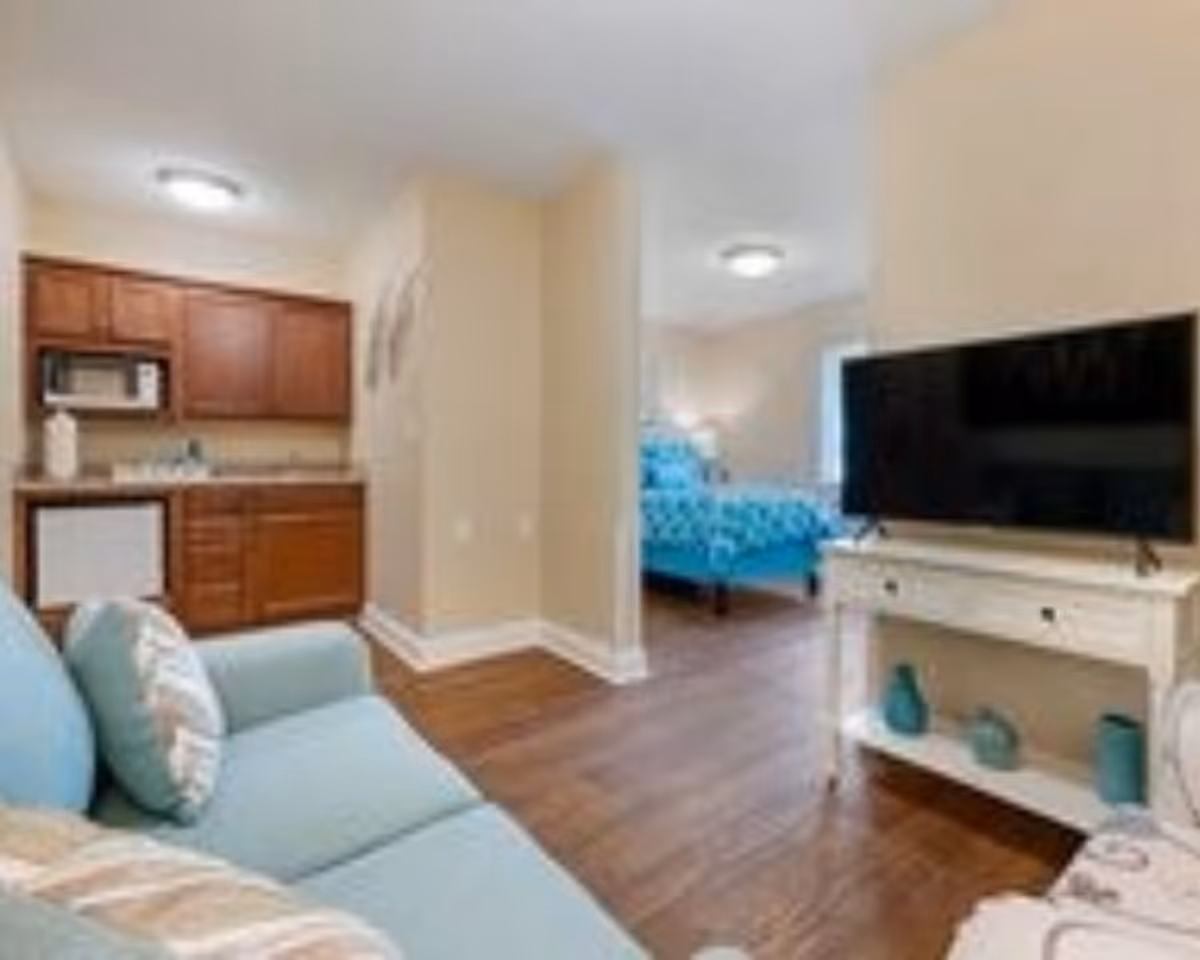 Studio-style interior showing a light-blue sofa, small kitchenette, TV on a console, and a bed visible through an open doorway.