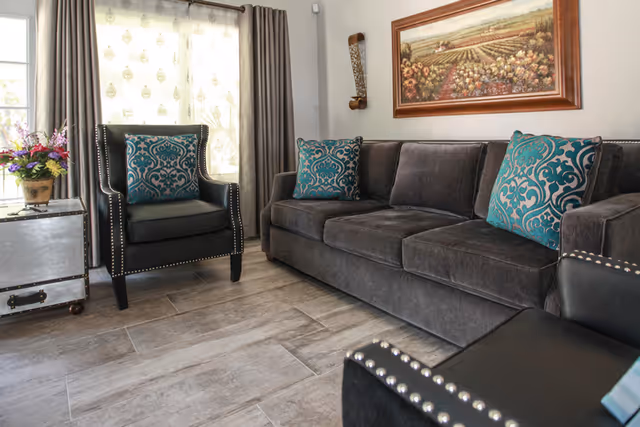 A cozy living room area featuring a dark gray sofa with three teal and gray patterned cushions, a matching armchair with a similar cushion, a small silver chest used as a side table with a flower arrangement on top, light gray wood-look tile flooring, a window with sheer and dark curtains, and a framed painting of a vineyard on the wall.