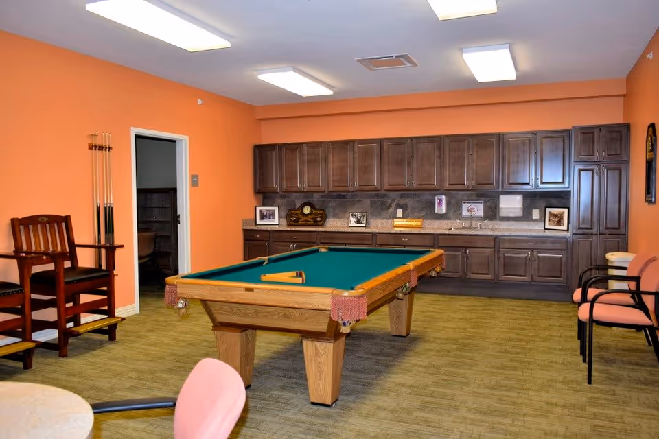 Interior room with an orange wall featuring a pool table in the center, a wooden chair with pool cues on the left, and a long countertop with dark wooden cabinets and framed pictures on the back wall. There are also pink cushioned chairs on the right side of the room.
