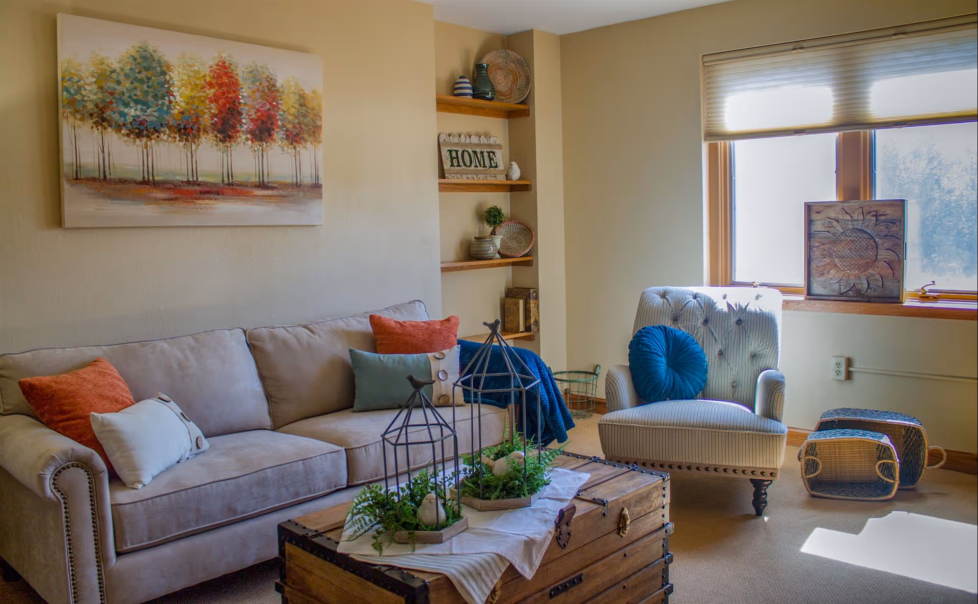 A cozy living room with a beige sofa adorned with colorful cushions, a wooden chest used as a coffee table with decorative plants and birdcages, a striped armchair with a blue round pillow, and a window with blinds letting in natural light. There are wooden shelves with decorative items and a painting of colorful trees on the wall.
