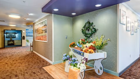 Interior hallway of a senior living facility with a decorative flower cart filled with colorful artificial flowers on the right side. The hallway has carpeted floors and walls adorned with framed pictures. The ceiling has recessed lighting and a purple accent panel.