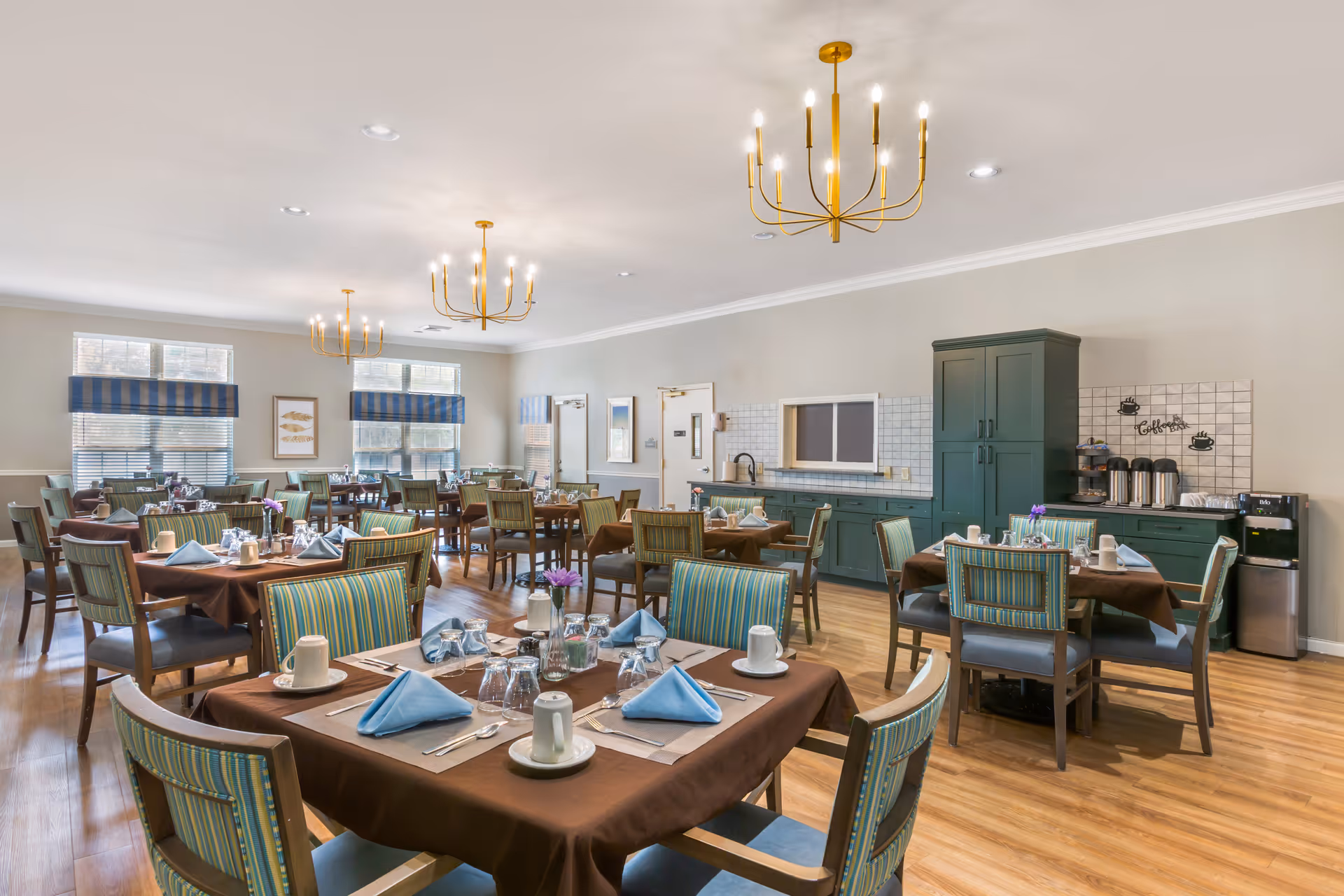 A bright dining room with multiple tables set for a meal. Each table has brown tablecloths, blue folded napkins, white cups, glasses, and silverware. The chairs have striped upholstery in shades of green and blue. The room features wooden flooring, large windows with blue and white striped valances, and three modern gold chandeliers hanging from the ceiling. Along one wall, there is a green cabinet area with a coffee station and a small sink.