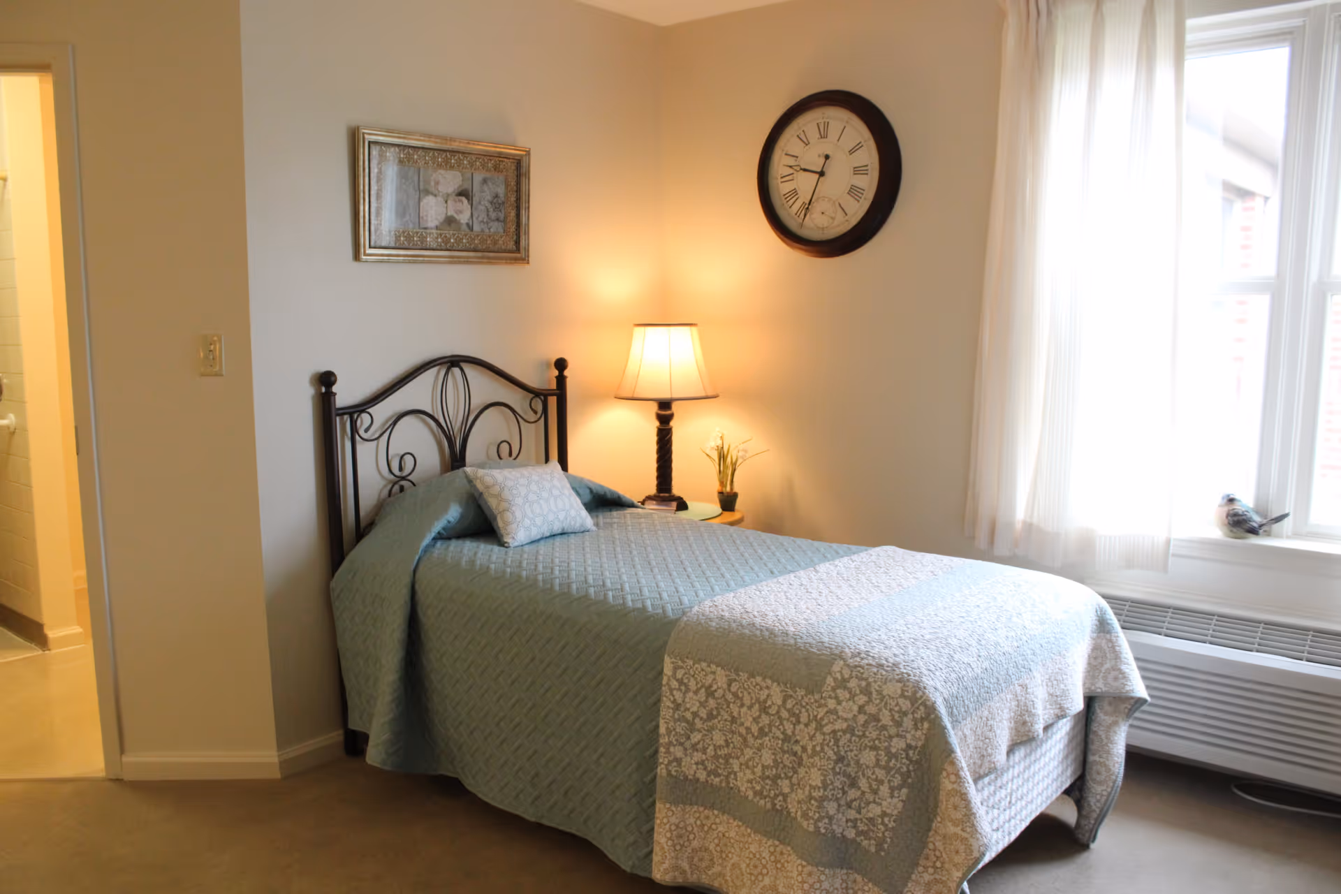 A cozy bedroom with a single bed covered in a light blue quilt and a decorative pillow. There is a bedside table with a lamp and a small plant. A large round clock hangs on the wall above the lamp, and a framed picture is on the adjacent wall. Natural light comes through a window with sheer white curtains.