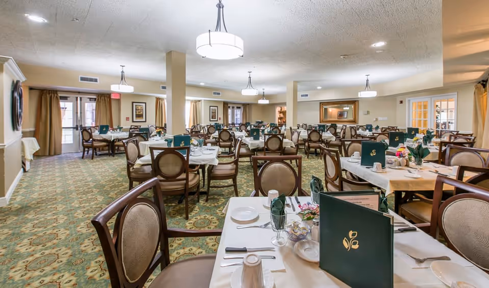 A spacious dining room with multiple tables set for a meal, each table covered with white tablecloths, green menus, and neatly arranged silverware and napkins. The room has patterned carpet, beige walls, several ceiling lights, and large windows with curtains allowing natural light to enter.