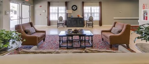 A senior living facility lobby seating area with sofas and chairs arranged around a coffee table on a patterned rug, with large windows and entry doors in the background.