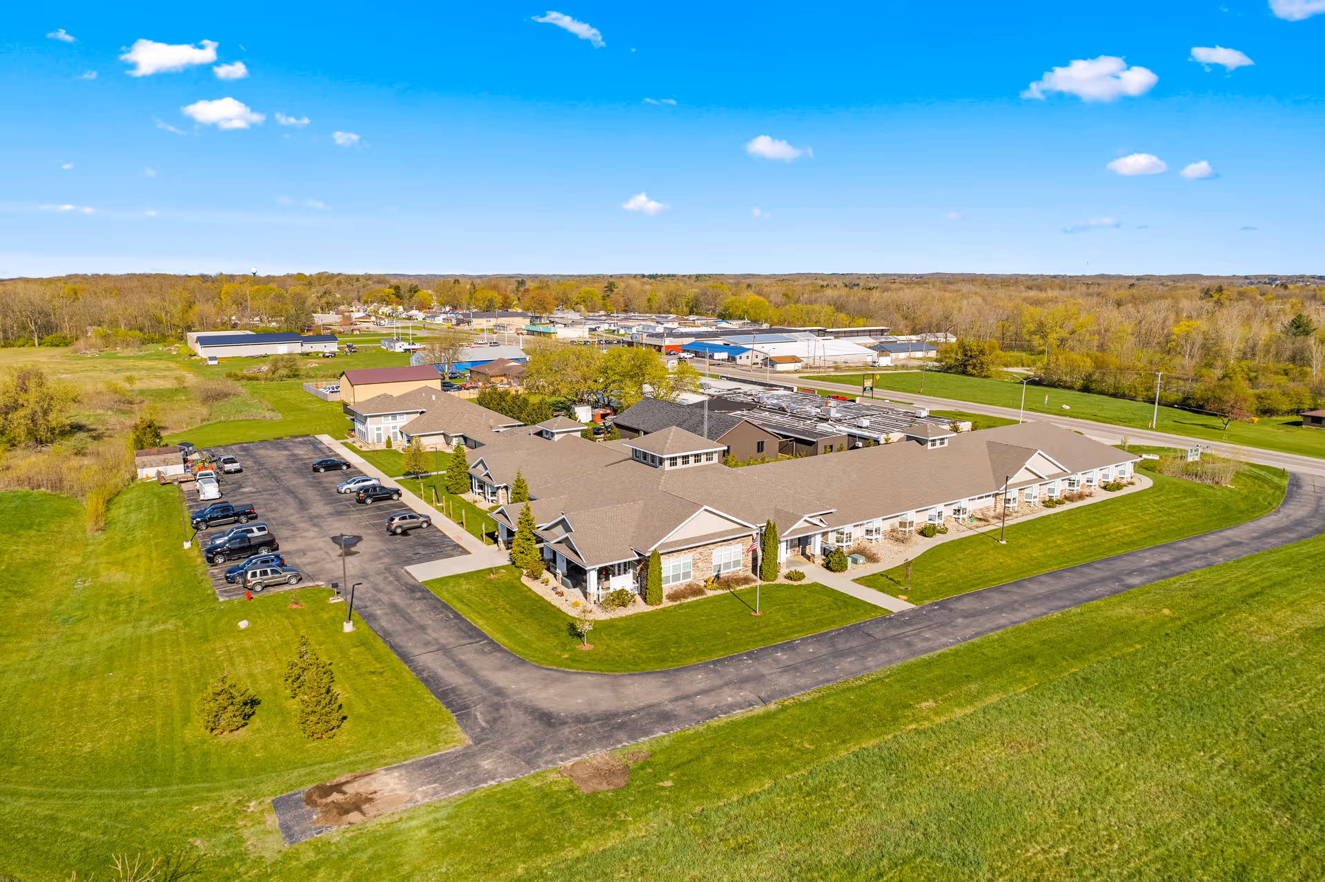 Aerial view of Woodland Terrace of Paw Paw Lake, showing a large single-story building with a brown roof surrounded by green lawns and a parking lot with several cars. The facility is located in a semi-rural area with trees and open fields in the background under a blue sky with scattered clouds.