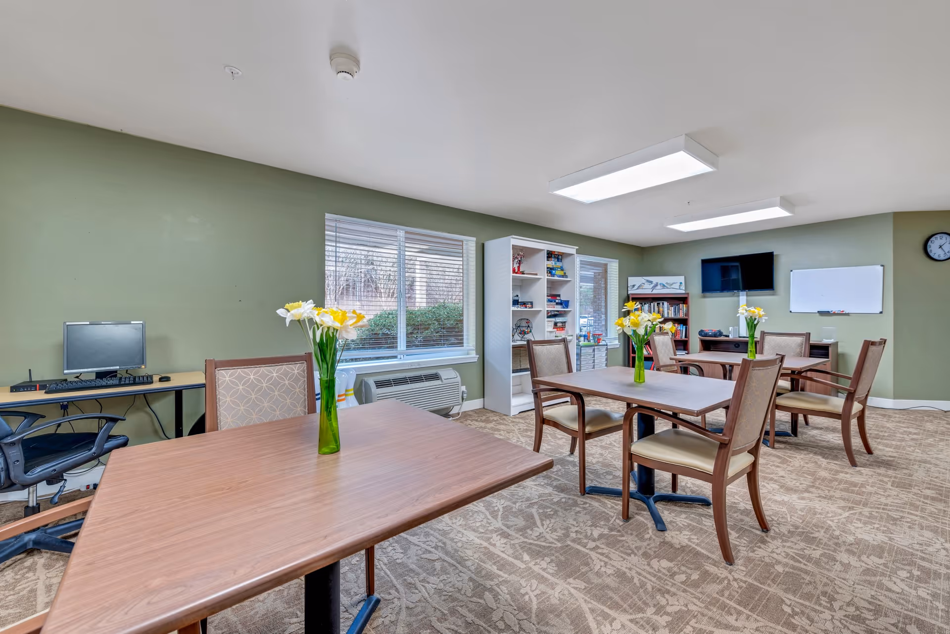 A well-lit activity room with green walls and beige patterned carpet. The room contains several wooden tables with chairs, each table decorated with a green vase holding yellow and white flowers. There is a computer workstation with a chair along one wall, two windows with blinds, a white bookshelf filled with board games and books, a wall-mounted TV, a whiteboard, and a clock on the wall.