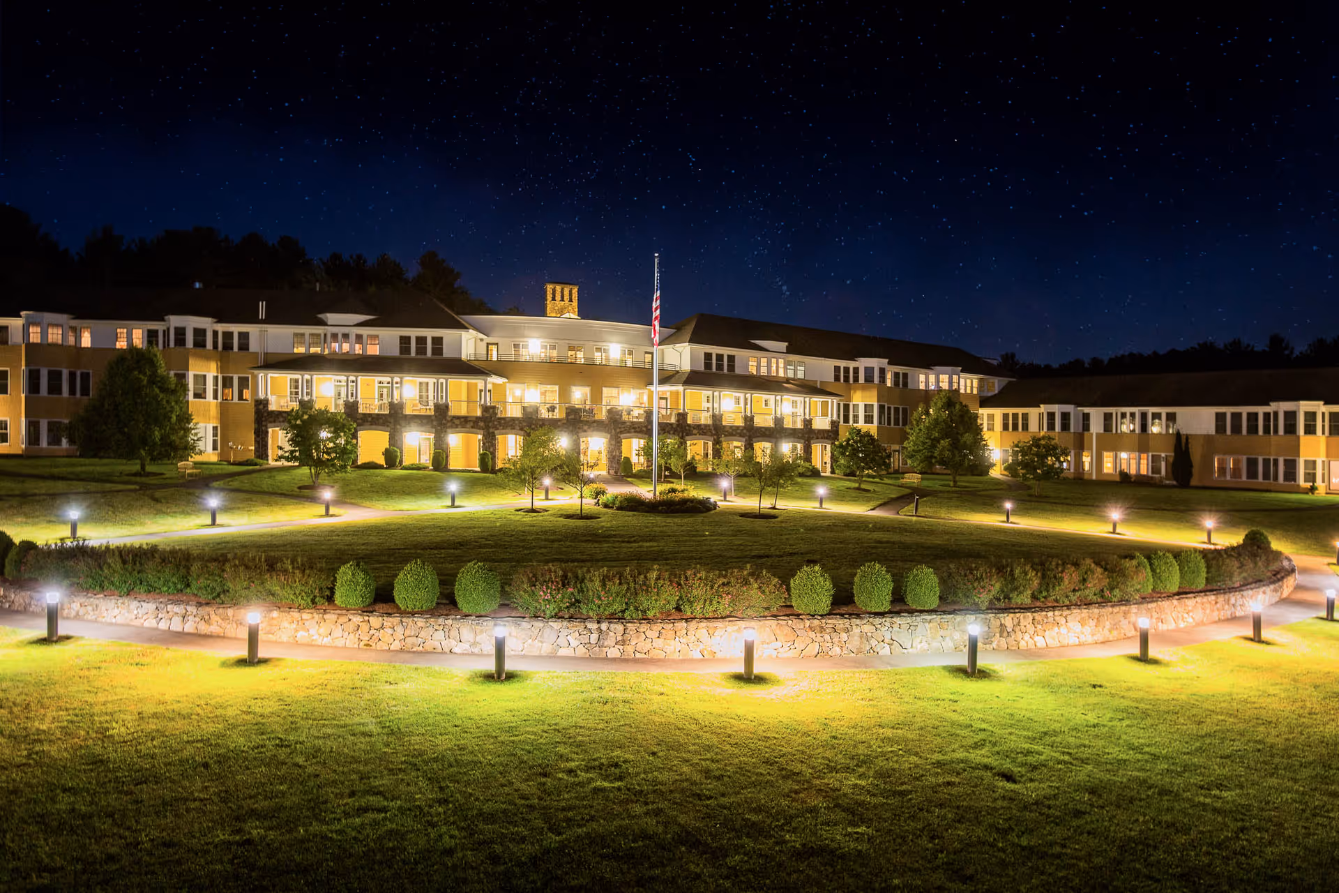 Nighttime view of Nichols Village's illuminated three-story building with a circular landscaped lawn and flagpole in front.